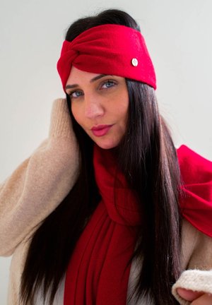 Fascia in maglia rossa con design attorcigliato, caratterizzata da un accento metallico. La texture morbida si abbina a una sciarpa rossa coordinata e a un maglione beige.