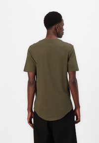 Tricou scurt cu mânecă, de culoare verde măslin, fabricat din bumbac, având un tiv curbat și un design simplu. Textură uniformă, fără modele sau accente vizibile.