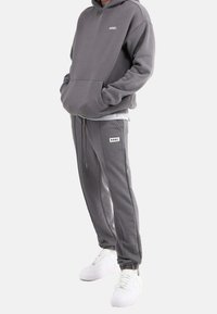Szara bluza z kapturem i dopasowane spodnie dresowe wykonane z miękkiego materiału, z przednią kieszenią i detalem z logo. Noszone z białymi trampkami.