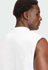 Valkoinen hihaton T-paita, jossa on kaareva kaula-aukko, sileä pinta ja näkyvät ompelusaumat, päällä henkilö, joka on hieman poispäin kääntyneenä.