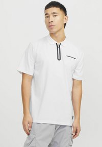 Hvid kortærmet polo shirt med sort lynlås-lukning, der har et subtilt logo på venstre side. Glat tekstur, afslappet pasform, enkel design.
