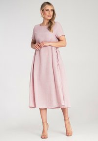 Rosa knielanges Kleid mit kurzen Ärmeln, runder Ausschnitt, gerafften Seitenschnüren und einem leichten, strukturierten Stoff. Getragen mit nudefarbigen Riemchenheels.