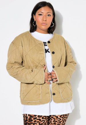 Persoon draagt een beige doorgestikte jas over een wit overhemd en een broek met luipaardprint, kijkt recht vooruit met handen bij de knopen van de jas.
