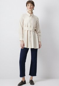 Krämfärgad skjorta med knytmidja, lång ärm och avslappnad passform, kombinerad med mörk denimjeans med fransar vid fållen och loafers.