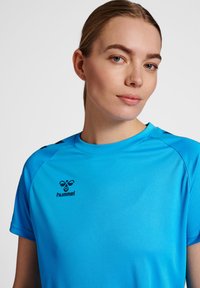 Blå sportstrøje med rund hals, korte ærmer og sorte accenter. Har "hummel" logoet på forsiden, lavet af glat stof.