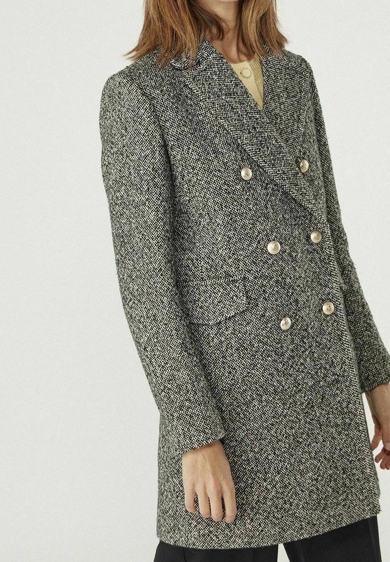 Femme portant un blazer gris en tweed à double boutonnage avec six boutons métalliques et des poches à rabat, associé à un pantalon noir.