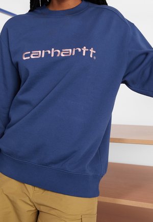 Person trägt ein marineblaues Carhartt-Sweatshirt mit hellrosa Logotext und beigen Cargohosen, steht in der Nähe von Holzmöbeln.