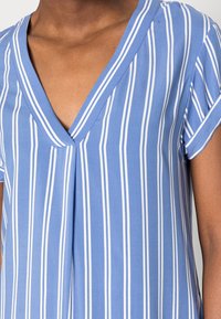 Blus i blå och vit randig design med V-ringning, korta ärmar och vertikala vita ränder. Slät textur med lätt och andningsbar tyg.