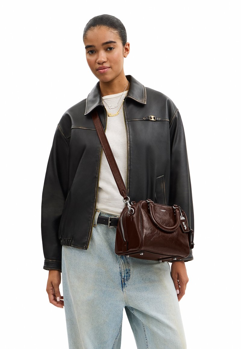 Jeune femme portant une veste en cuir noire, une chemise blanche, un jean bleu clair, et un sac à main en cuir marron avec une bandoulière.