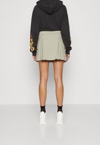 Plisovaná sukně s vysokým pasem ve světle olivově zelené barvě, doplněná černou mikinou. Viditelné jsou bílé tenisky a černé ponožky s volánkovým okrajem.