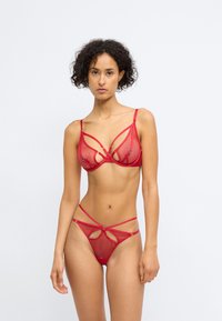 Conjunto de lingerie em rede vermelha com um soutien triangular com um design de alças intricado e uma tanga correspondente com recortes em forma de chave e alças ajustáveis.