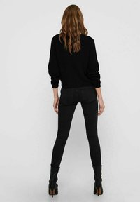 Schwarzer gerippter Pullover mit langen Ärmeln, kombiniert mit schwarzen Skinny-Jeans und schwarzen High-Heels. Haare, die bis zur Mitte des Rückens fallen.