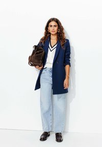 Blazer oversize bleu marine, top en tricot blanc à col en v, jeans à jambes larges bleu clair et mocassins brillants marron foncé. Sac en suède marron à la main.