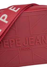 Röd crossbody-väska med präglad logotyp, texturerad yta och en vävd rem med varumärkesnamn i krämfärg.