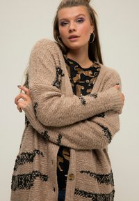 Beige Strickjacke mit schwarzen Tiermustern und gerippten Bündchen. Verfügt über einen Knopfverschluss und einen lockeren Schnitt, der über einem schwarzen Oberteil mit goldener Druckgrafik getragen wird.