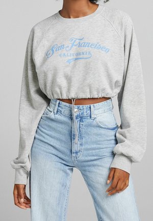 Grauer, kurz geschnittener Pullover mit blauem Schriftzug „San Francisco California“, getragen mit hoch taillierter hellblauer Jeans an einer Person.