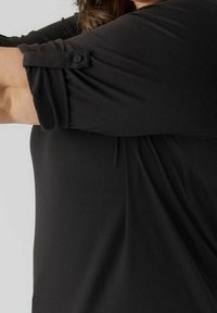 Maglietta nera a maniche corte con orli arrotolati, tessuto morbido, superficie liscia e dettagli con bottoni. Design semplice con una vestibilità rilassata.