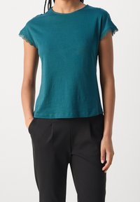 Teal, kortärmad T-shirt med scallop-lace kanter, mjukt bomullsmaterial, avslappnad passform, kombinerad med svarta byxor.
