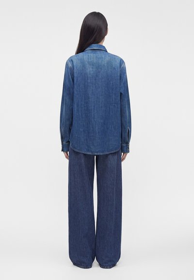 Femme aux longs cheveux lisses portant une chemise en jean ample à manches longues et un jean large bleu foncé, debout, de dos.
