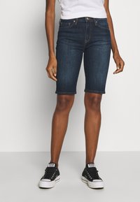 Bermudas de mezclilla oscura con un diseño ajustado, cintura de media altura y dobladillo enrollado. Combinadas con zapatillas negras y una camiseta blanca.