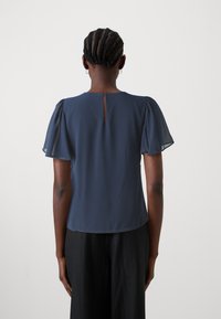 Mörkblå blus med korta genomskinliga ärmar, en detalj med nyckelhål i ryggen, och ett mjukt texturerat tyg. Bärs med svarta byxor.
