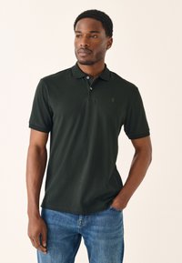 Ciemnozielona koszulka polo z miękkiej tkaniny, z trzema guzikami i haftowanym akcentem na piersi, w zestawie z niebieskimi dżinsami.