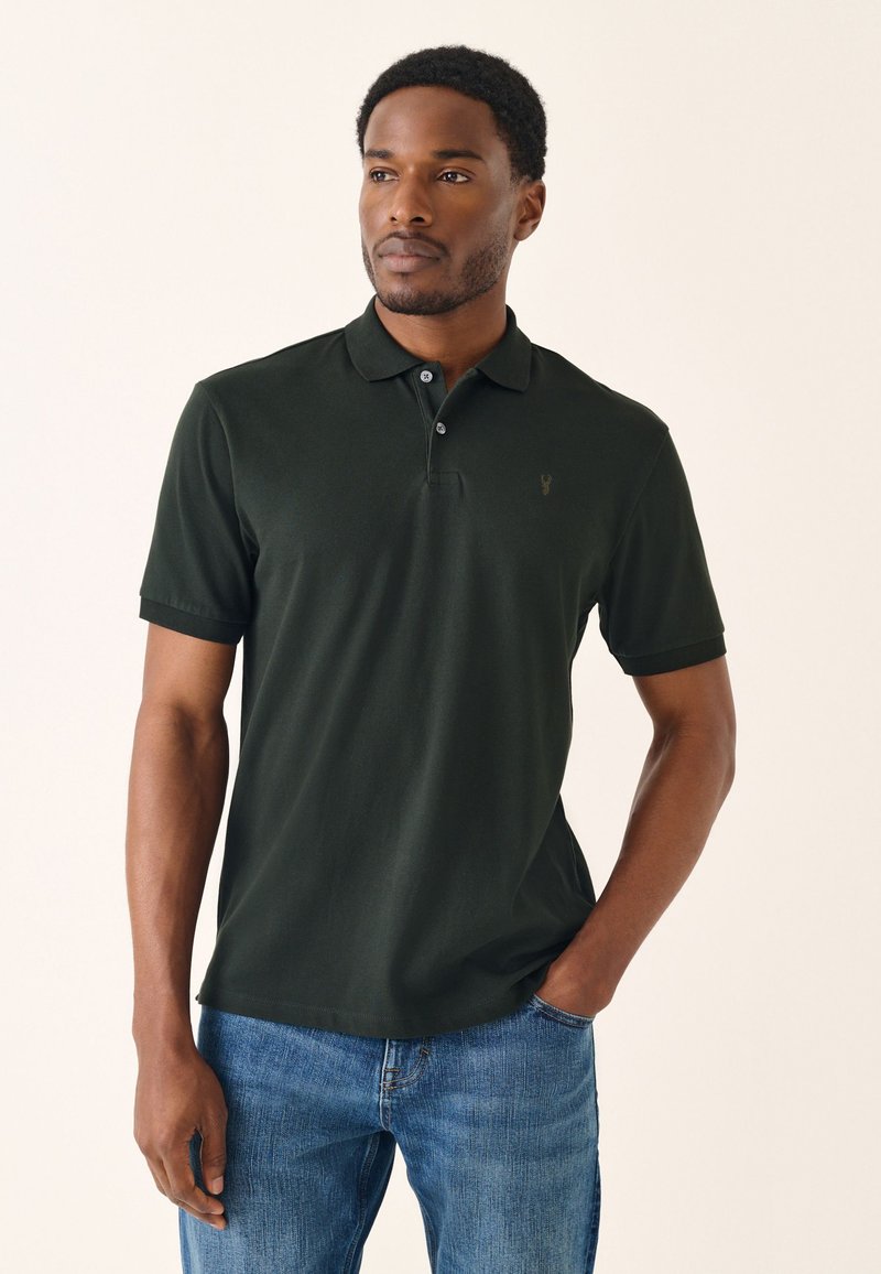 Ciemnozielona koszulka polo z miękkiej tkaniny, z trzema guzikami i haftowanym akcentem na piersi, w zestawie z niebieskimi dżinsami.