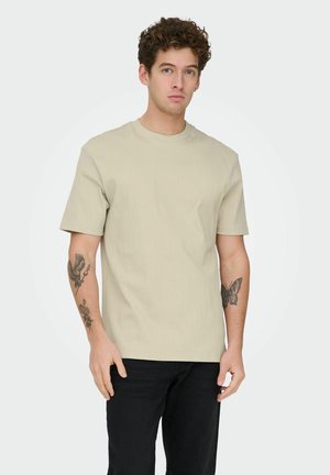 Młody mężczyzna z kręconymi włosami, ubrany w beżową koszulę z krótkim rękawem i czarne spodnie, pokazujący tatuaże na przedramionach na białym tle.