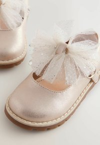 Metallisch pinkfarbene Kinderschuhe mit einer gewellten Öffnung, dekoriert mit einer großen weißen Tüllschleife und Glitzerelementen. Gummisohle.