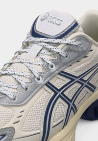 Κοντινό πλάνο λευκού και ναυτικού μπλε παπουτσιού τρεξίματος Asics με λεπτομέρειες δεσίματος κορδονιών και διαπνέον επάνω μέρος από δίχτυ.