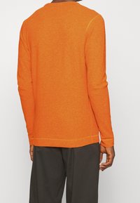 Oranssi neulottu pitkähihainen pusero, jossa on teksturoitu pinta, pyöreä pääntie ja sivuhalkiot helmassa, yhdistettynä tummiin housuihin.