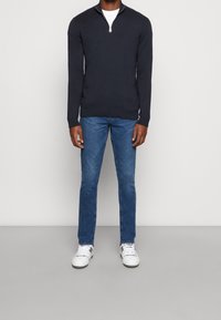 Pullover navy con collo a zip abbinato a jeans blu e sneaker bianche. Il pullover presenta un bordo e polsini a costine, realizzato in materiale leggero.
