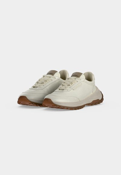 Ζευγάρι off-white αθλητικά παπούτσια με χοντρές καφέ σόλες και μπεζ λογότυπο στη γλώσσα, εμφανίζονται σε ελαφρώς γωνιακή θέση σε απλό φόντο.