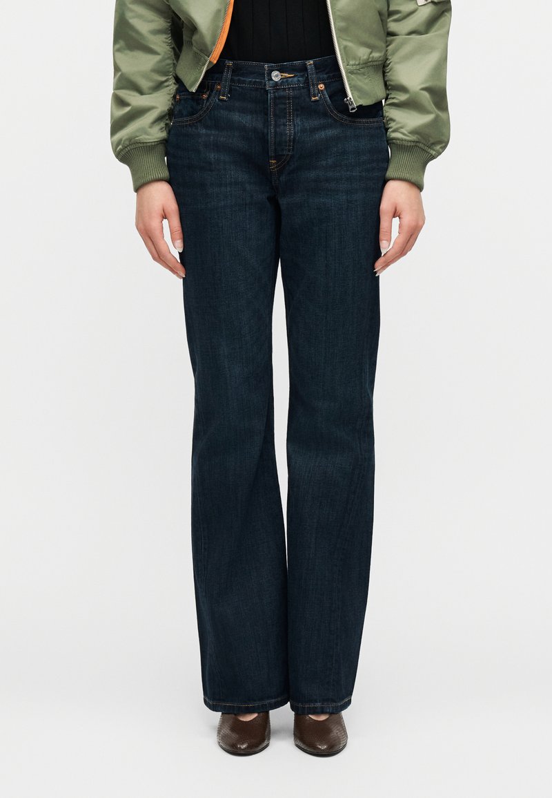 Γυναίκα που φοράει ψηλόμεσο σκούρο μπλε τζιν παντελόνι με φαρδιά μπατζάκια, συνδυασμένο με πράσινο μπουφάν τύπου bomber και καφέ μυτερά παπούτσια, στέκεται όρθια.