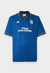 Mėlyna futbolo marškinėliai su juodu apykakle ir rankovių kraštais, balti "TIKROS SPALVOS" tekstas, Umbro logotipas ir Mancunian Way FC herbas ant krūtinės.