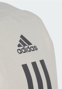 T-shirt de sport gris clair avec logo Adidas gris foncé et trois bandes. Le tissu a une finition texturée. Gros plan sur la zone de l'épaule.
