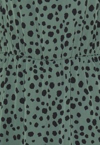 Tissu vert avec une texture lisse, présentant un motif de points noirs de tailles variées, et un design plissé à la taille pour plus de détails.