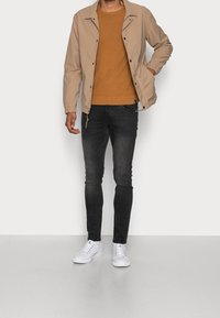 Man som bär en beige jacka över en brun tröja, svarta tajta jeans och vita sneakers, stående mot en enkel ljus bakgrund.