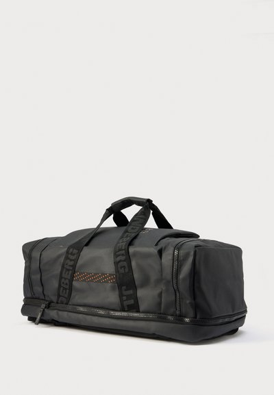 Sac de sport noir souple avec deux poignées, poche zippée latérale et sangles de marque, présenté sur un fond gris clair.