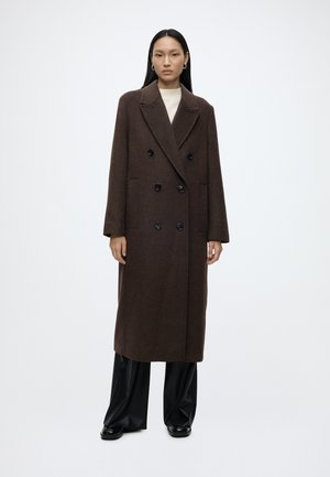 Femme debout portant un long manteau croisé marron foncé, un pantalon noir à jambes larges et des chaussures noires sur un fond blanc uni.