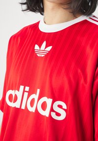 Rood stoffen sporttruitje met witte accenten, gestreept ontwerp en een wit "adidas" logo met daarboven een driebladig embleem.