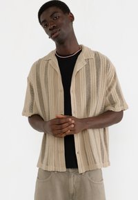 Strikbluse med korte ærmer i beige med lodrette striber, letvægtigt stof, afslappet pasform, knaplukning foran, båret over en sort T-shirt.