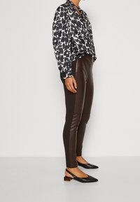 Persona vestida con blusa floral en blanco y negro, legging de imitación cuero marrón oscuro y zapatos de tacón bajo negros tipo slingback, de pie de lado.
