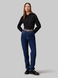 Čierna skrátená mikina s logom Calvin Klein, spárovaná s vysokými modrými džínami a čiernymi robustnými topánkami, na sivom pozadí.