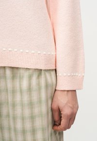 Χέρι που ακουμπά στο πλάι, φοράει ανοιχτό ροζ πουλόβερ με λευκή διακόσμηση με τελείες και μπεζ καρό φούστα με πράσινες ρίγες.