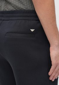 Pantaloni sport negri cu textură netedă, având un buzunar pe spate, talie elastică și un mic accent logo argintiu pe partea din spate.