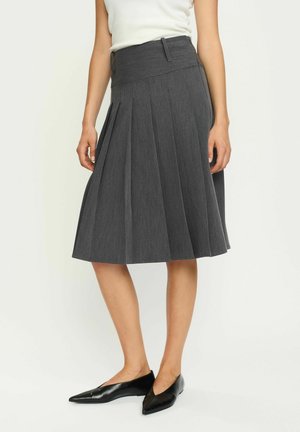 Faltbarer grauer Rock mit hoher Taille, aus strukturiertem Stoff gefertigt. Getragen mit schwarzen spitzen Ballerinas. Strukturiertes Design in Knielänge.