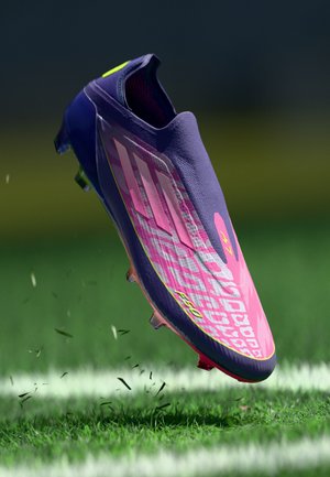 Cristiană de fotbal violet cu un design roz și galben, suprafață texturată și cuțe translucide. Este suspendată deasupra unui teren de iarbă.