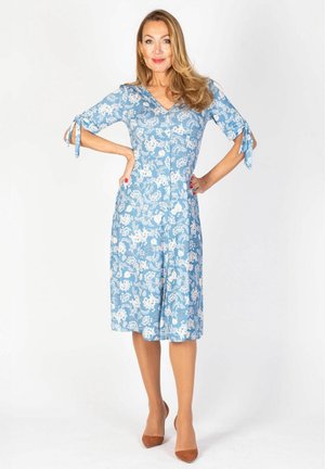 Kobieta stojąca z rękami na biodrach, ubrana w niebieską sukienkę w kwiaty do kolan i brązowe szpilki na jednolitym białym tle.