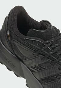 Czarne buty sportowe z teksturowaną cholewką wykonaną z materiałów syntetycznych, wzmocnionym noskiem oraz siateczkową podszewką i amortyzowaną podeszwą.